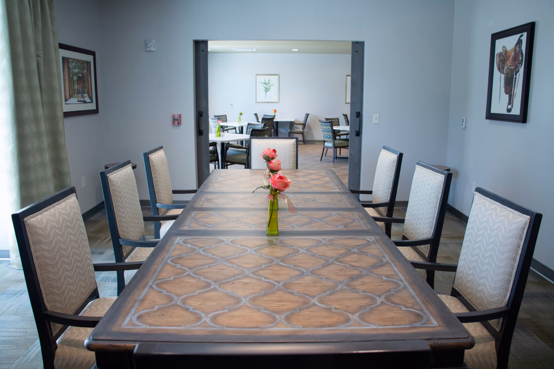 Long wooden dining table with patterned top and chairs around it, a small vase of pink flowers centered and an open doorway to another dining area.