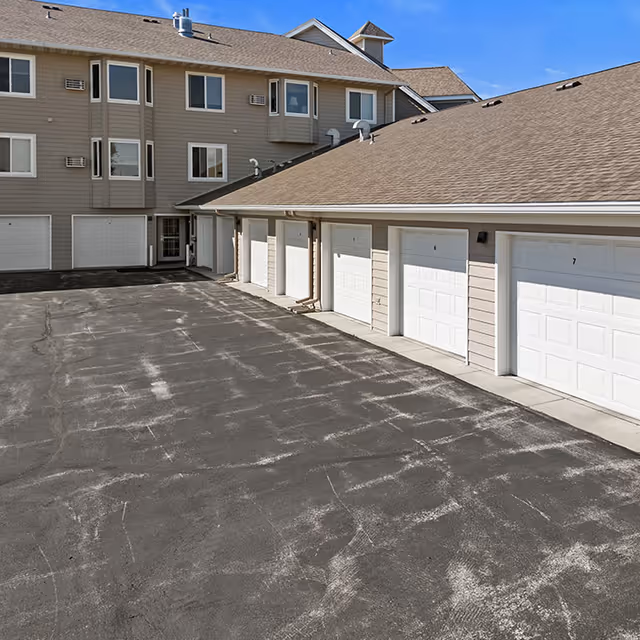 Exterior view of a senior living facility showing a row of closed white garage doors attached to a beige building with multiple windows under a clear blue sky.