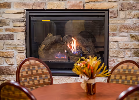Round table with chairs and a floral centerpiece in front of a stone-surrounded gas fireplace.
