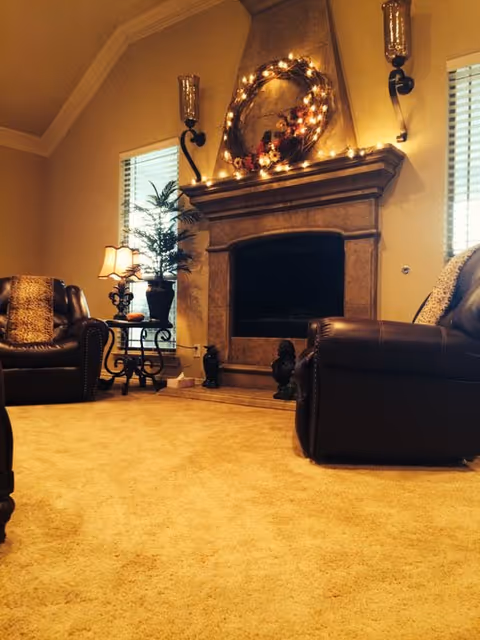 Cozy living room with beige carpet, two dark leather armchairs with cushions, a stone fireplace decorated with a lighted wreath, two windows with blinds, a small side table with a lamp and a potted plant.