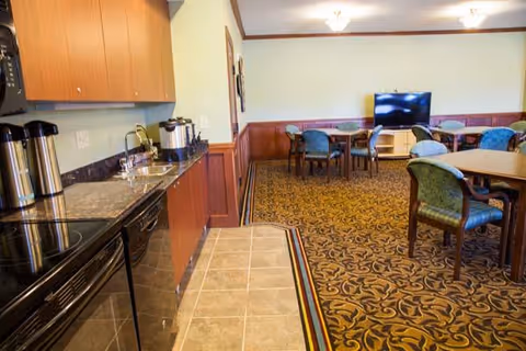 Interior view of a senior living facility common area featuring a kitchenette with a countertop, sink, coffee urns, and cabinets on the left. The room has a patterned carpet with several round tables and chairs arranged for seating. A flat-screen TV is mounted on a stand against the far wall.