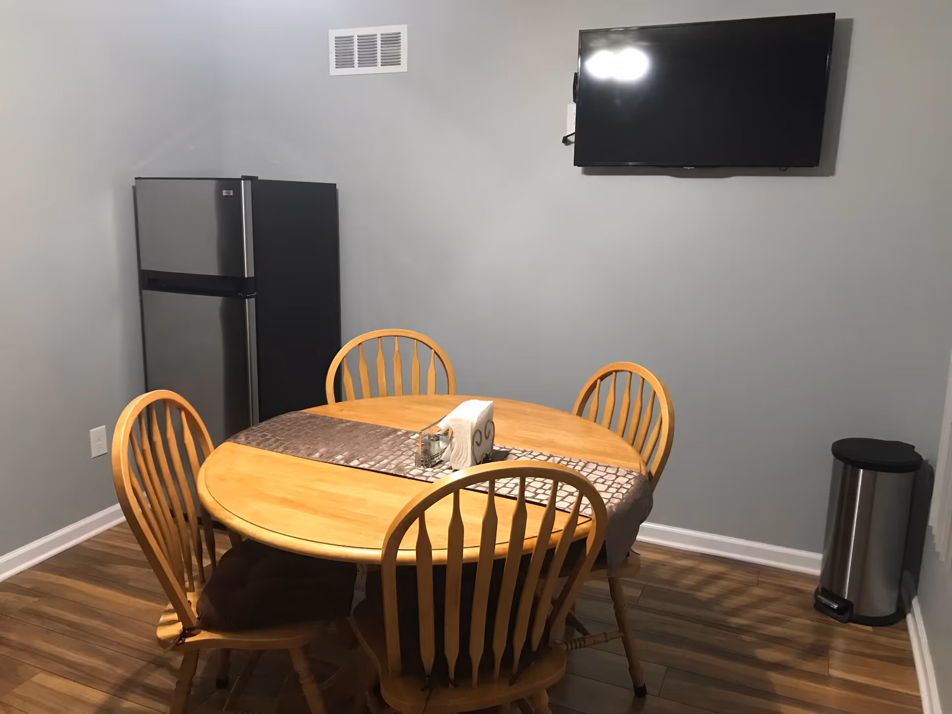 Small dining area with a round wooden table and four chairs, a wall-mounted TV, and a refrigerator in the corner.