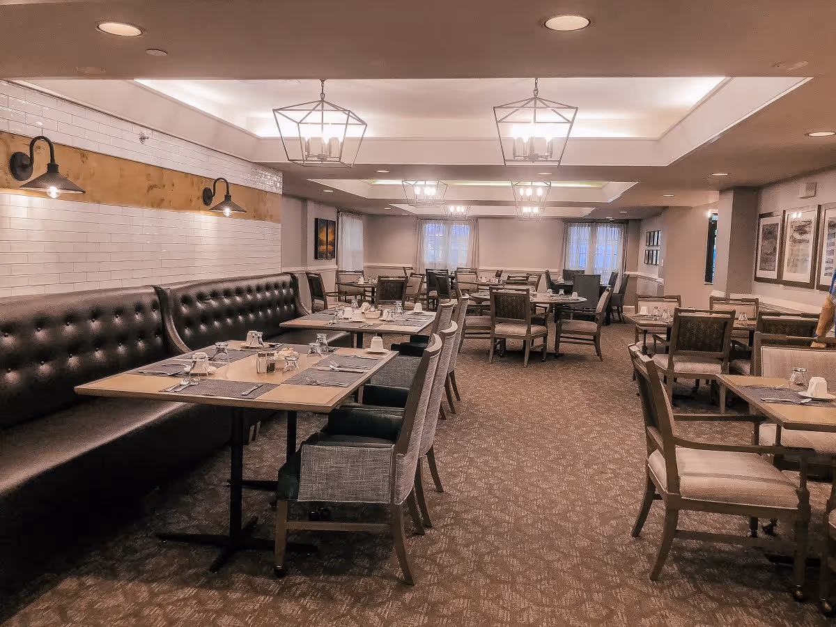A spacious dining room with multiple tables and chairs arranged neatly. The tables are set with placemats, glasses, and utensils. There is a long black cushioned bench along one wall with wall-mounted lights above it. The ceiling has recessed lighting and decorative hanging light fixtures. Windows with curtains are visible at the far end of the room.
