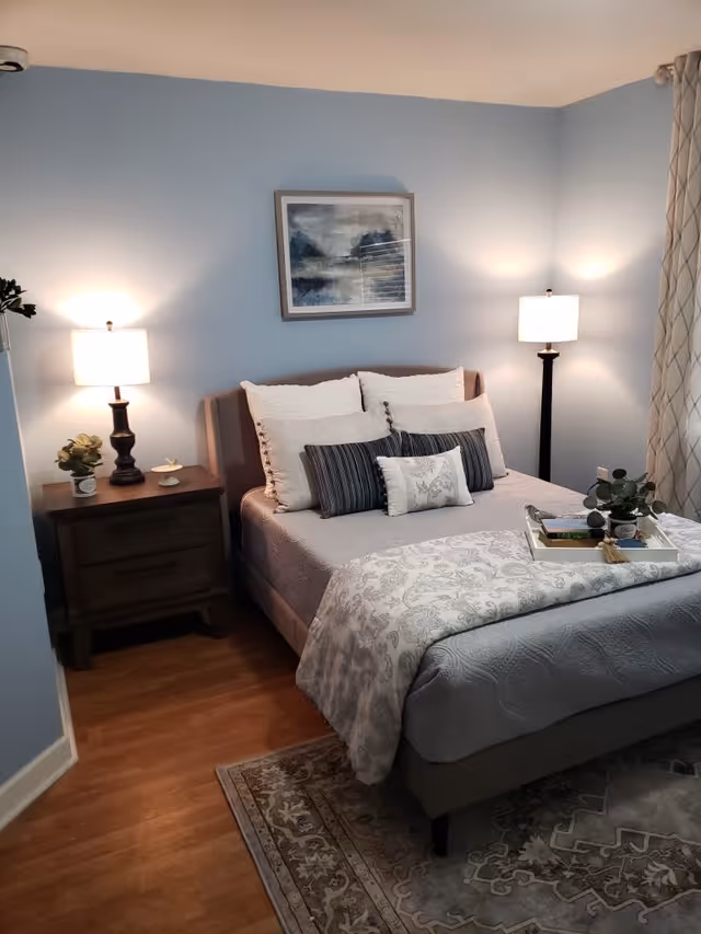 A neatly made bed with multiple pillows and a patterned blanket in a bedroom with light blue walls. There is a nightstand with a lamp and a small plant on the left side of the bed, and a tall floor lamp on the right side. A framed abstract painting hangs above the bed, and a tray with books and a small plant is placed on the bed. The floor is wooden with a patterned rug partially visible.