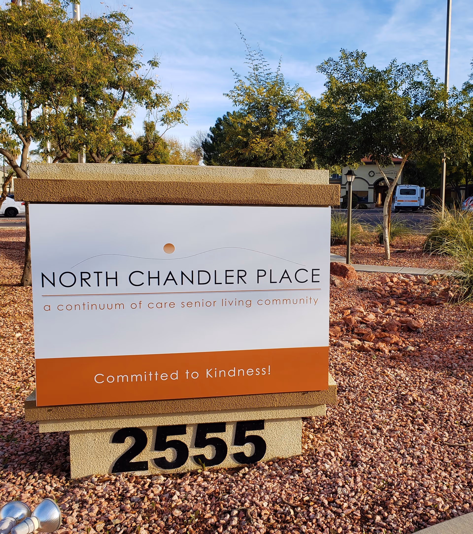 Outdoor sign for North Chandler Place, a continuum of care senior living community, with the slogan 'Committed to Kindness!' and the address number 2555. The sign is surrounded by landscaping with rocks and trees, and a building and parked vehicles are visible in the background.
