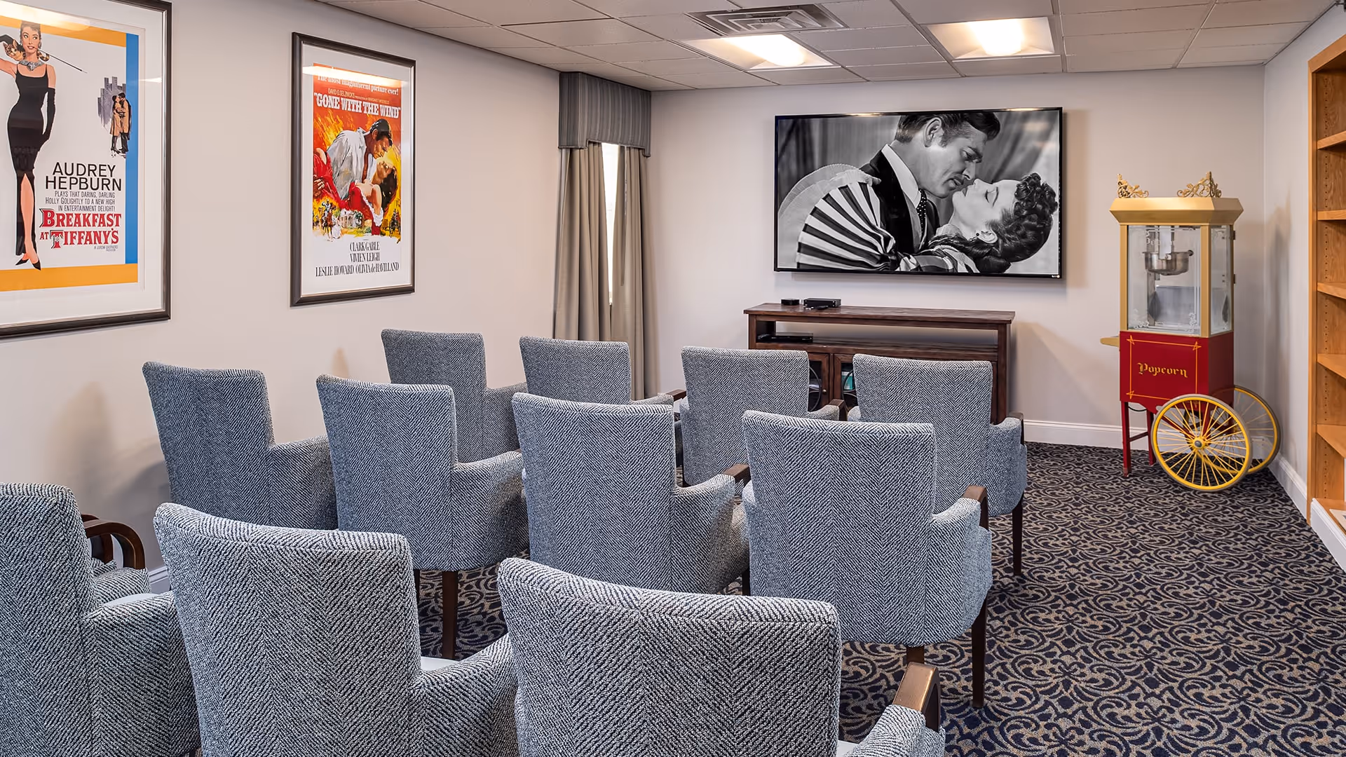 A small movie screening room with two rows of gray upholstered chairs facing a large TV screen displaying a black and white romantic movie scene. The room has patterned carpet, framed classic movie posters on the walls, a popcorn machine on the right side, and a wooden media console beneath the TV.