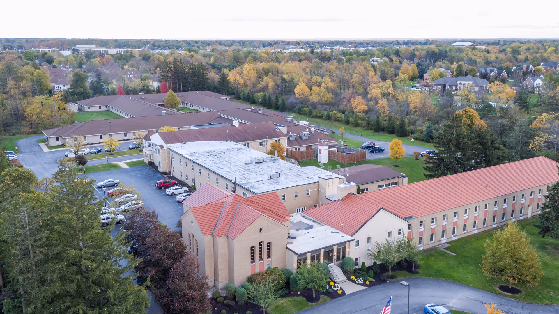 Aerial view of Brompton Heights of Williamsville senior living facility surrounded by trees with autumn foliage. The building has multiple wings with red and white roofs, parking lots with cars, and landscaped greenery around the entrance.