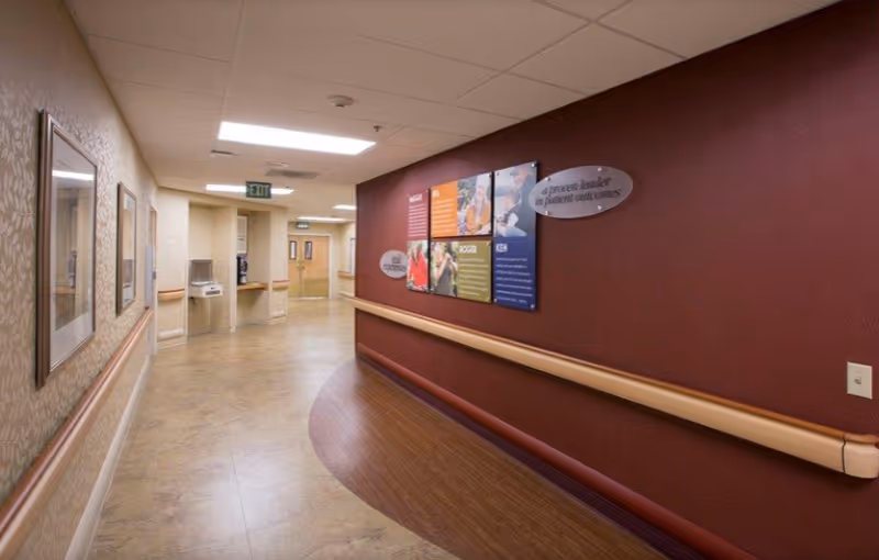 Well-lit curved interior hallway of a senior care facility with wooden handrails, framed artwork, and informational wall signs.