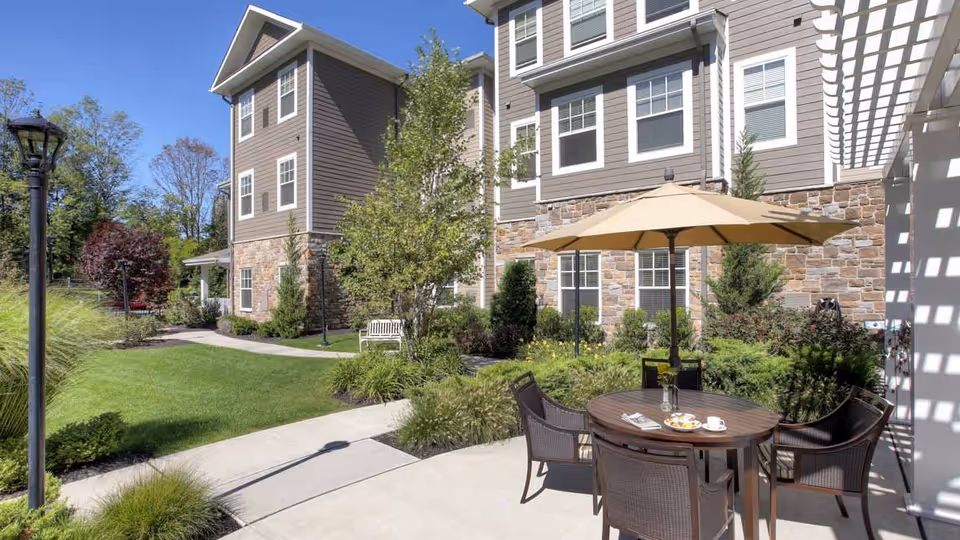 Outdoor patio area at Arbor Terrace Roseland featuring a round wooden table with four wicker chairs and a large beige umbrella. The patio is surrounded by green shrubs and plants, with a well-maintained lawn and a multi-story building with stone and siding exterior in the background under a clear blue sky.