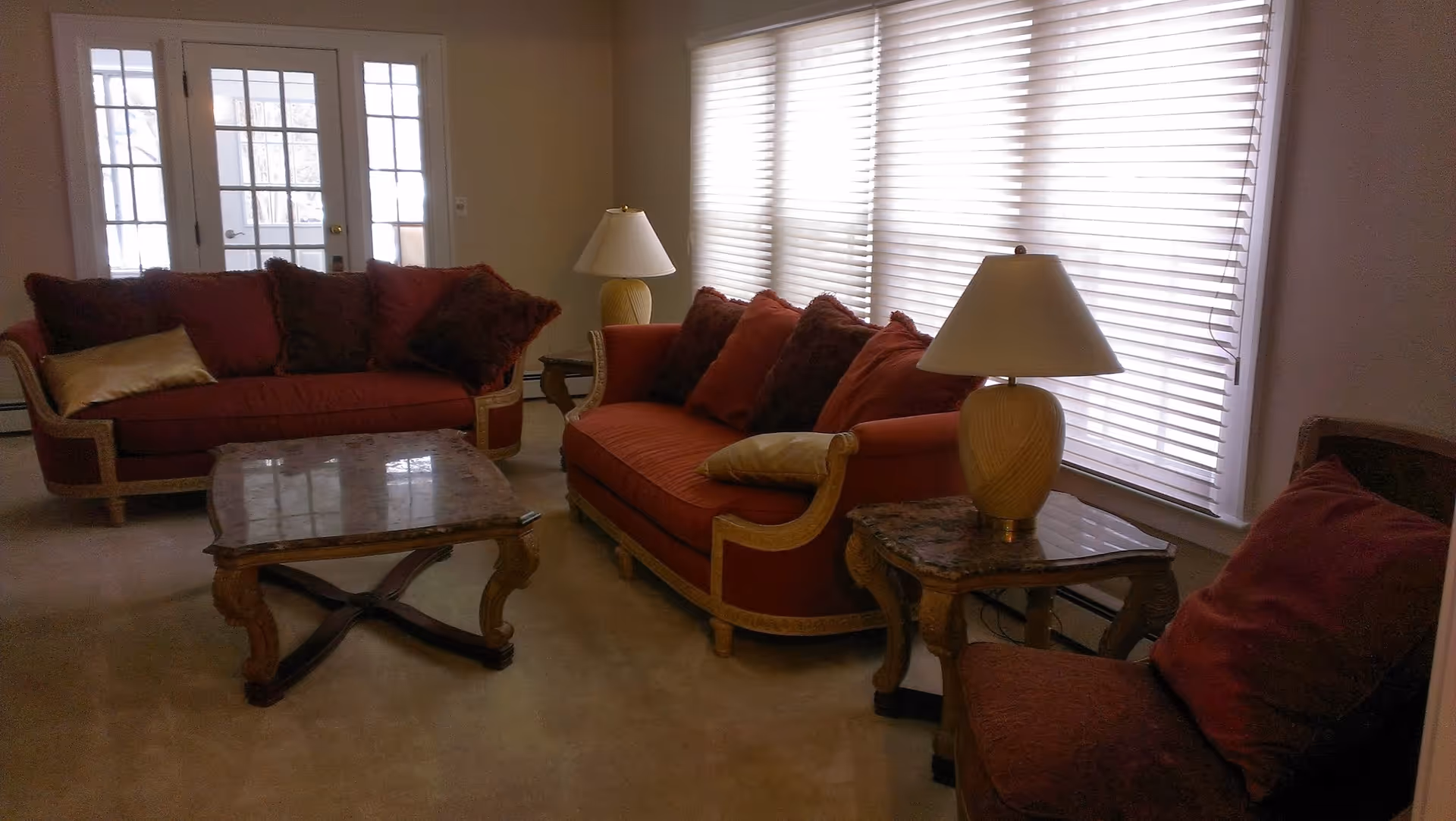 A living room with two red sofas and a matching armchair, all with decorative pillows. There are two wooden side tables with lamps on either side of one sofa, and a wooden coffee table in the center. Large windows with white blinds cover one wall, and a set of French doors is visible in the background.