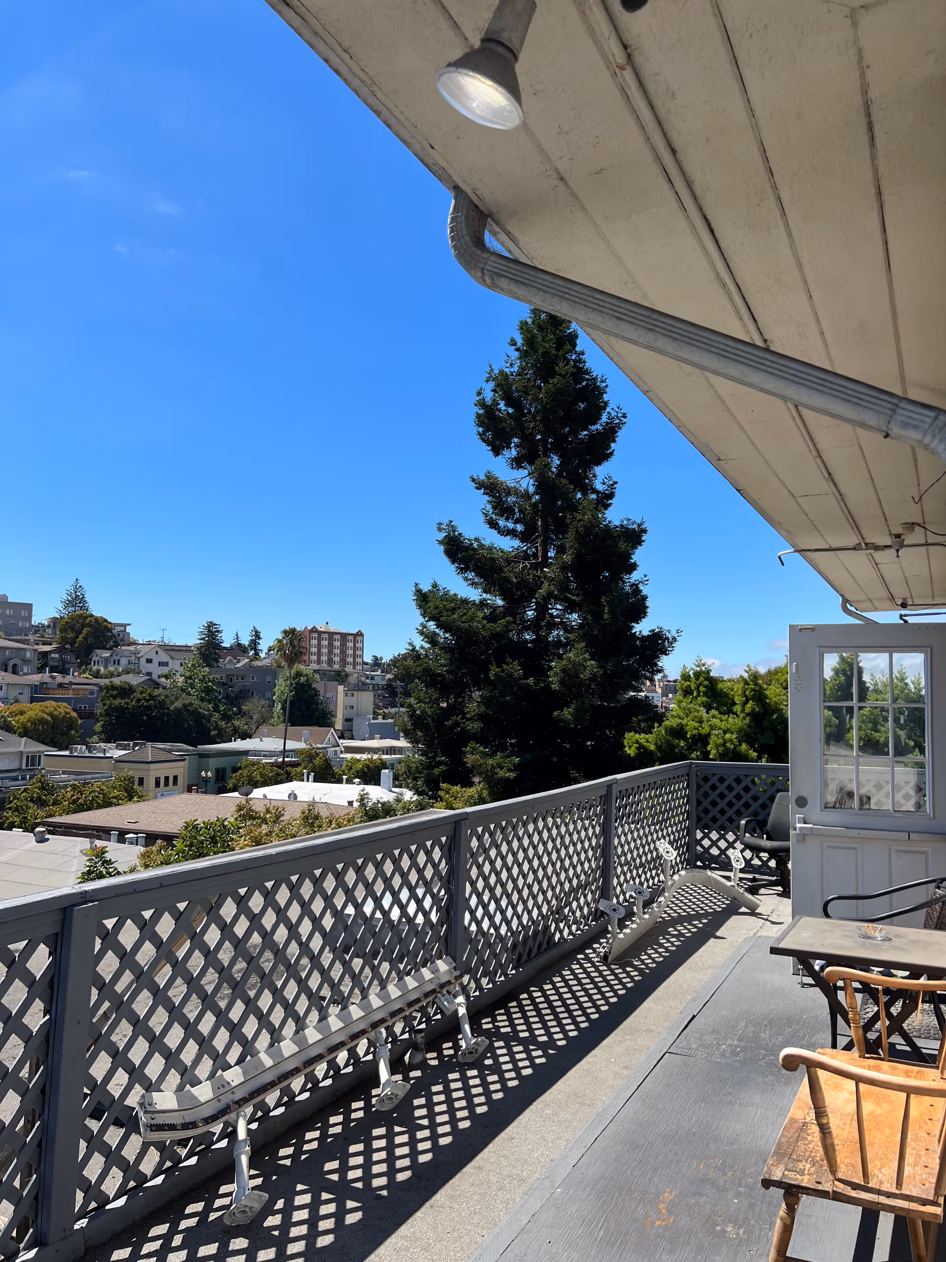 Outdoor balcony area with lattice railing, wooden chairs, a small table, and a view of nearby buildings and trees under a clear blue sky.
