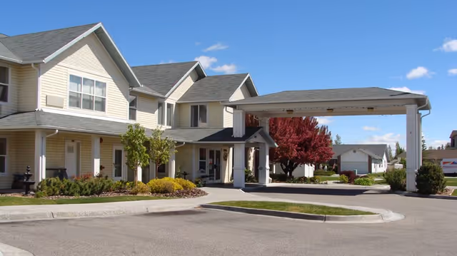 Exterior view of a two-story assisted living facility with beige siding and a covered entrance driveway. There are landscaped bushes and trees, including a tree with red leaves, under a clear blue sky.