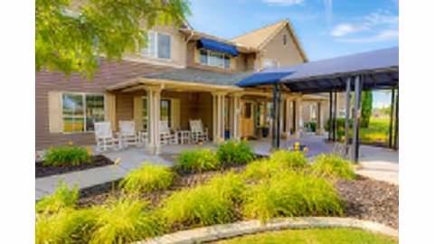 Exterior view of a senior living facility with a covered entrance, beige siding, blue awnings, and a landscaped garden with green shrubs and a tree in front.