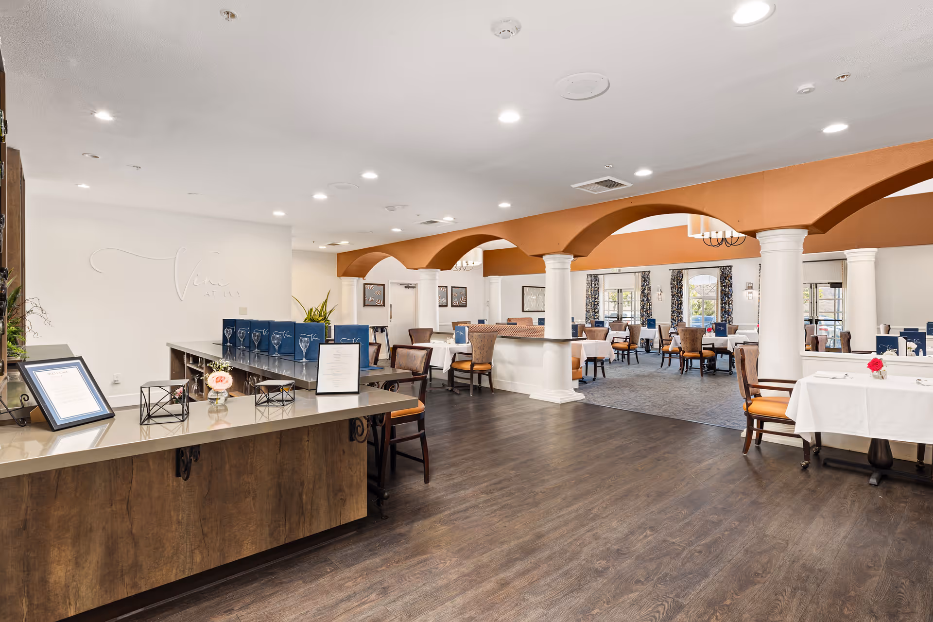Bright, spacious dining room with arched beams and columns, a reception counter in the foreground, and tables set with white tablecloths.