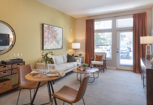 A furnished living room with a sofa, round coffee table, small dining table with two chairs, and a glass door with curtains leading outside.