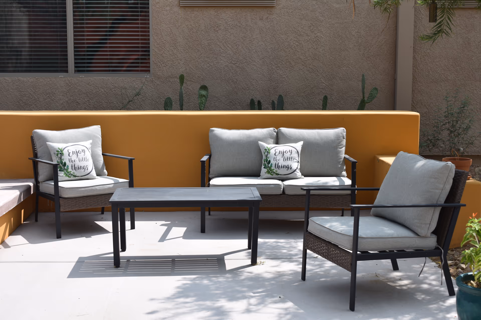 Outdoor patio area with cushioned seating including a loveseat and two armchairs around a black metal coffee table. The loveseat and one armchair have decorative pillows with the text 'Enjoy the little things'. The seating is placed against a mustard yellow wall with some potted plants and a window in the background.