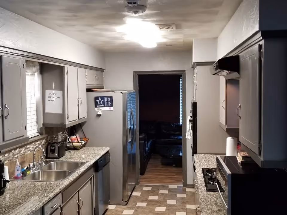 A narrow kitchen with gray cabinets and granite countertops on both sides. The left side features a double sink, a toaster, and a dishwasher, while the right side has a stove with a range hood and a microwave. A stainless steel refrigerator is at the end of the kitchen, with a doorway leading to a dimly lit living room with dark furniture. The floor has a patterned tile design, and there is a ceiling light fixture.