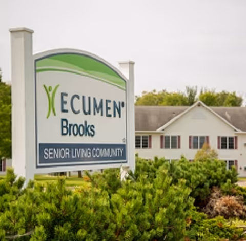 A yard sign reading "Ecumen Brooks Senior Living Community" in front of shrubs with a two-story residential building behind it.