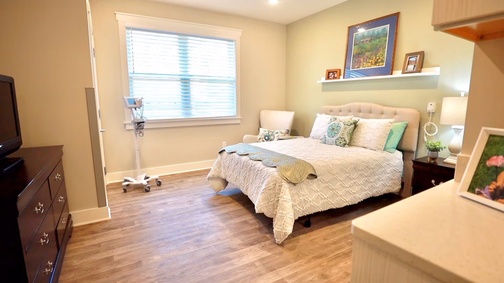 Furnished bedroom with a bed, armchair, dresser, lamp, and large window in an assisted living facility.