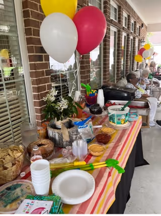 A festive outdoor buffet table set up with various snacks, dips, and disposable plates and cups. The table is covered with a striped tablecloth and decorated with white, yellow, and red balloons. Several elderly people are seated in the background along a brick wall with windows.