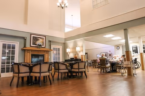 A spacious common area in a senior living facility with wooden flooring, a fireplace with a framed picture above it, and several chairs arranged around tables. In the background, a group of elderly people are seated at a table, engaged in conversation. The room has high ceilings with hanging light fixtures and a soft, neutral color palette.