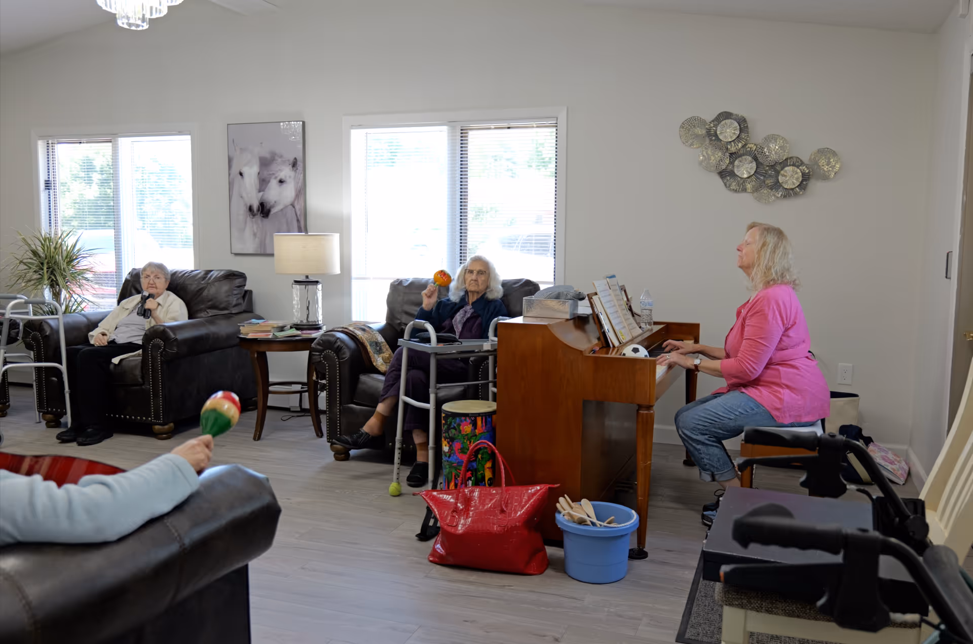 A group of elderly women in a bright living room at Meadow View Assisted Living & Memory Care. One woman in a pink shirt is playing a wooden piano, while two other women sit in leather armchairs holding maracas. The room has large windows with blinds, a side table with a lamp and books, and decorative wall art including a painting of two white horses and a metal wall sculpture.