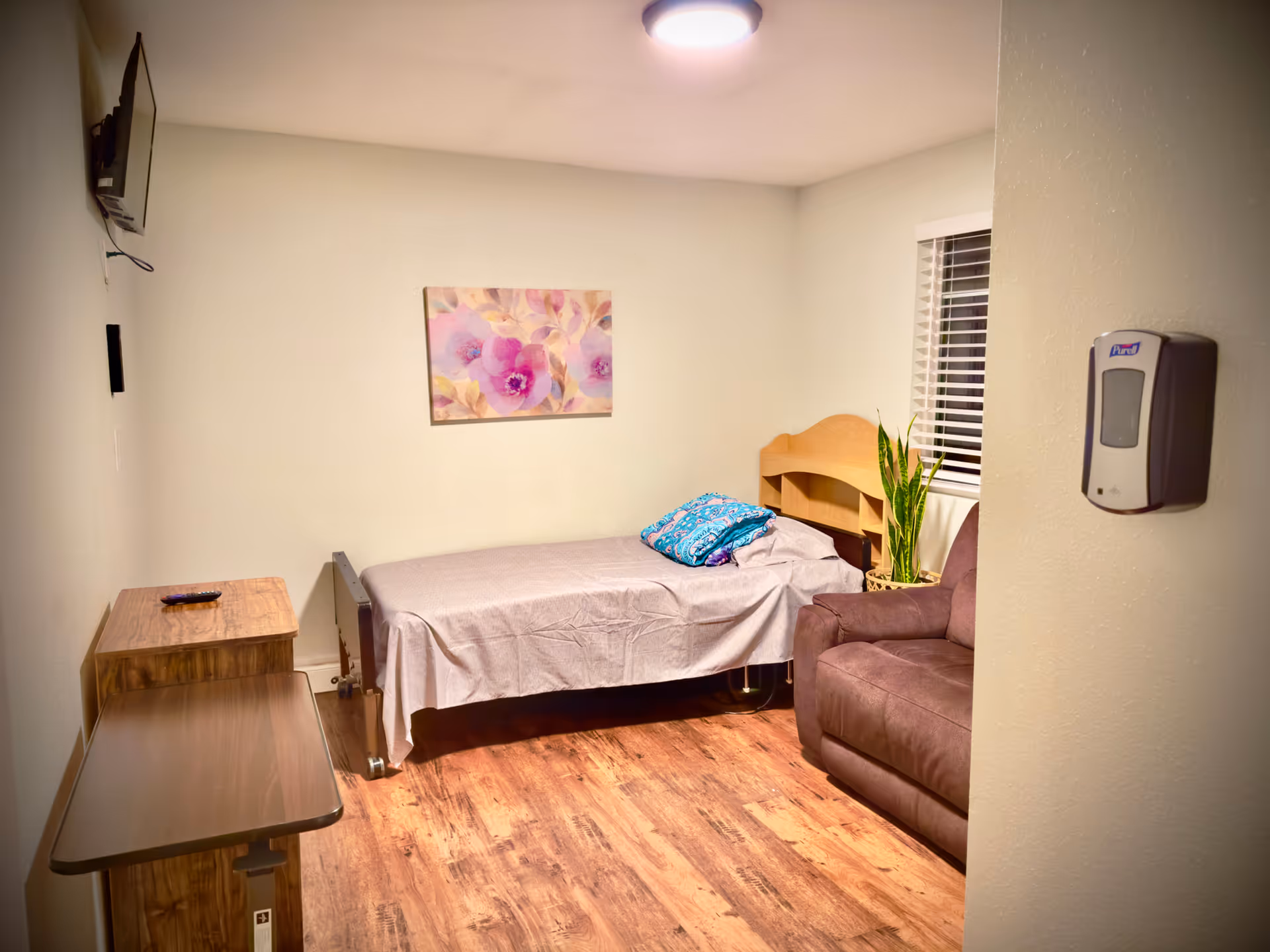 A small, cozy bedroom with a single bed covered with a light gray sheet and a folded blue blanket on top. There is a wooden nightstand with a remote control on it, a brown armchair, a potted plant near the window with white blinds, and a floral painting on the wall. A wall-mounted TV is visible on the left side, and a hand sanitizer dispenser is mounted on the right wall near the entrance.