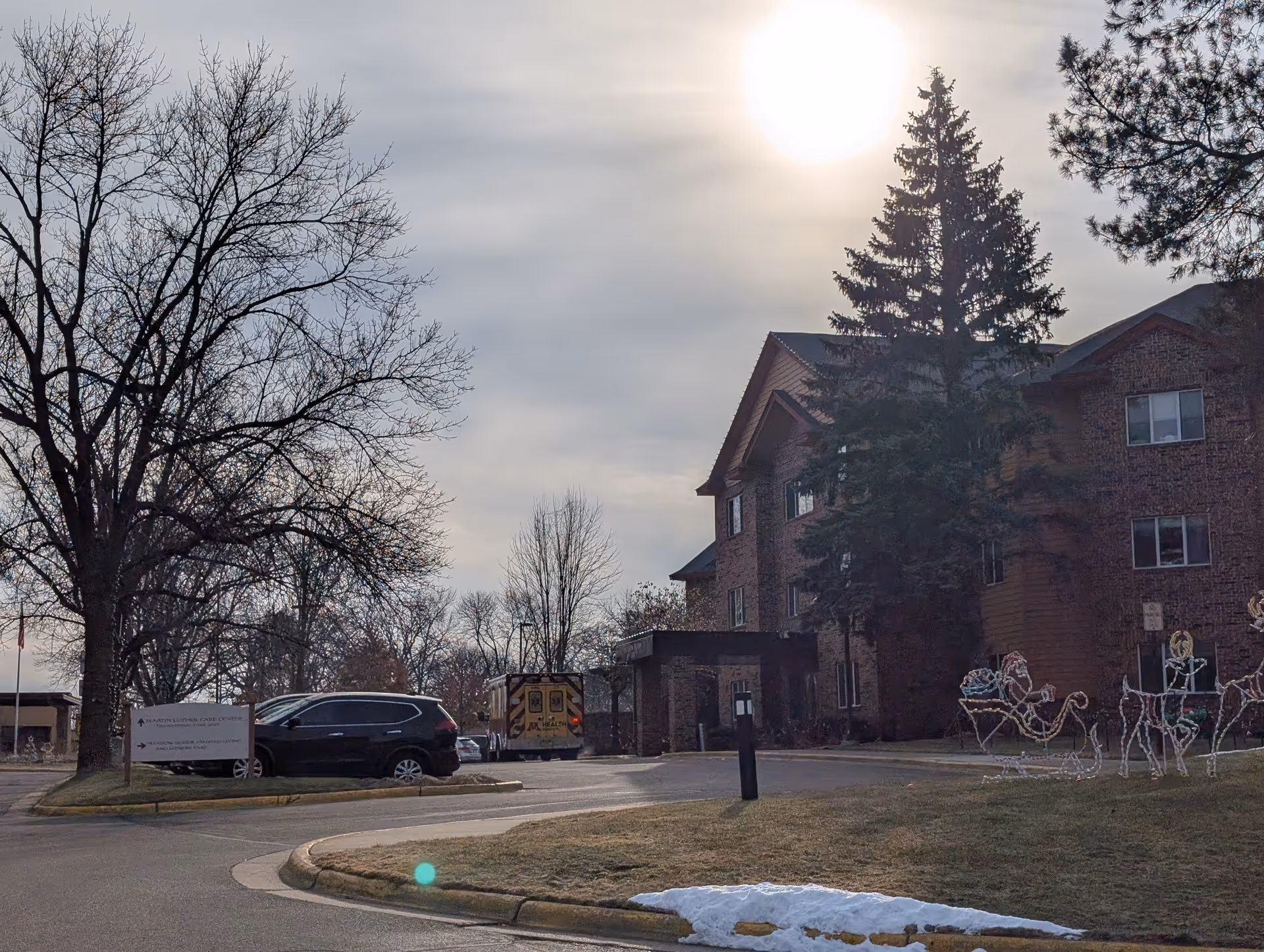 Exterior view of Martin Luther Campus building with a large tree and some holiday light decorations shaped like a sleigh and reindeer on the lawn. There are a few parked vehicles including an ambulance near the entrance. The sky is cloudy with the sun shining through.