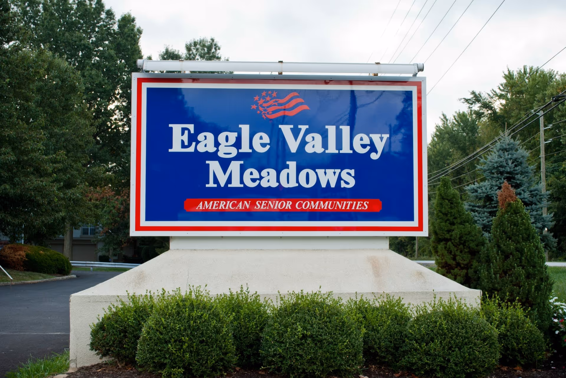A large outdoor sign for Eagle Valley Meadows, an American senior community, with a blue background, white and red text, and a small American flag graphic at the top. The sign is surrounded by greenery and trees.