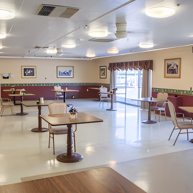 A spacious dining room with several small square tables and chairs arranged throughout. The room has a polished floor, beige walls with a maroon and green border, framed artwork on the walls, and a large sliding glass door with curtains letting in natural light.