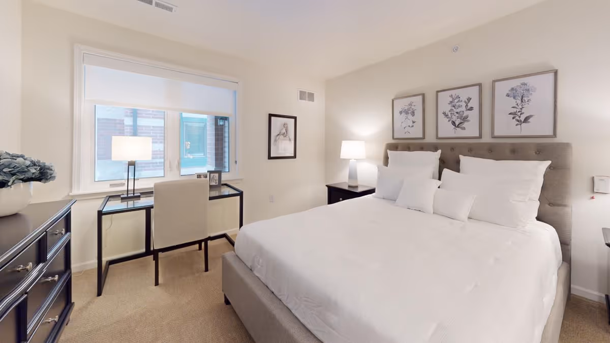 Bright furnished bedroom with a large bed, bedside tables, framed art, and a desk by the window.