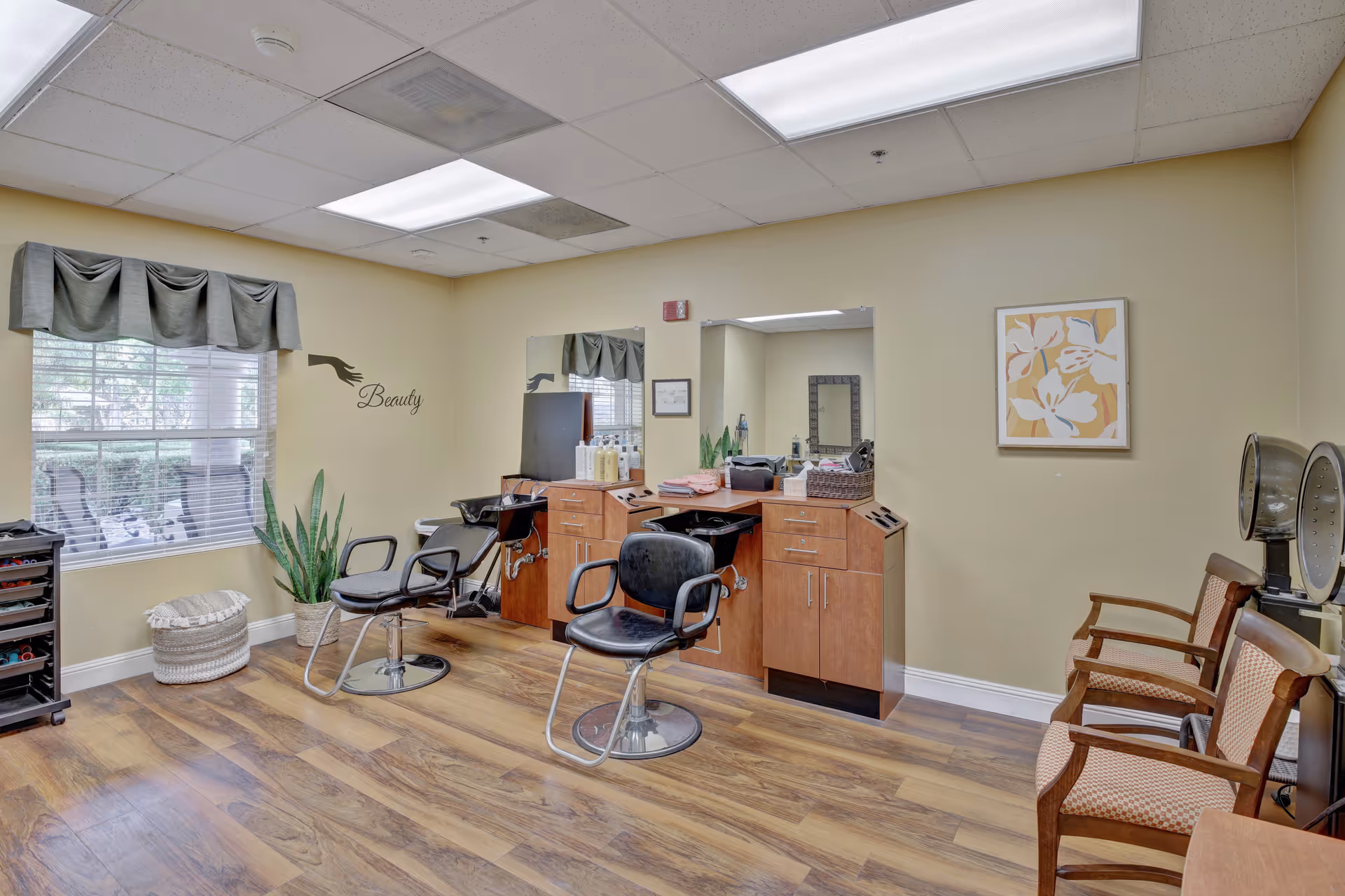 Interior of a beauty salon room in a senior living facility with two black salon chairs in front of wooden cabinets and mirrors. There is a window with a valance, a potted plant, a small ottoman, and a wall decoration that says 'Beauty'. On the right side, there are two wooden chairs with patterned cushions and two hair dryers.