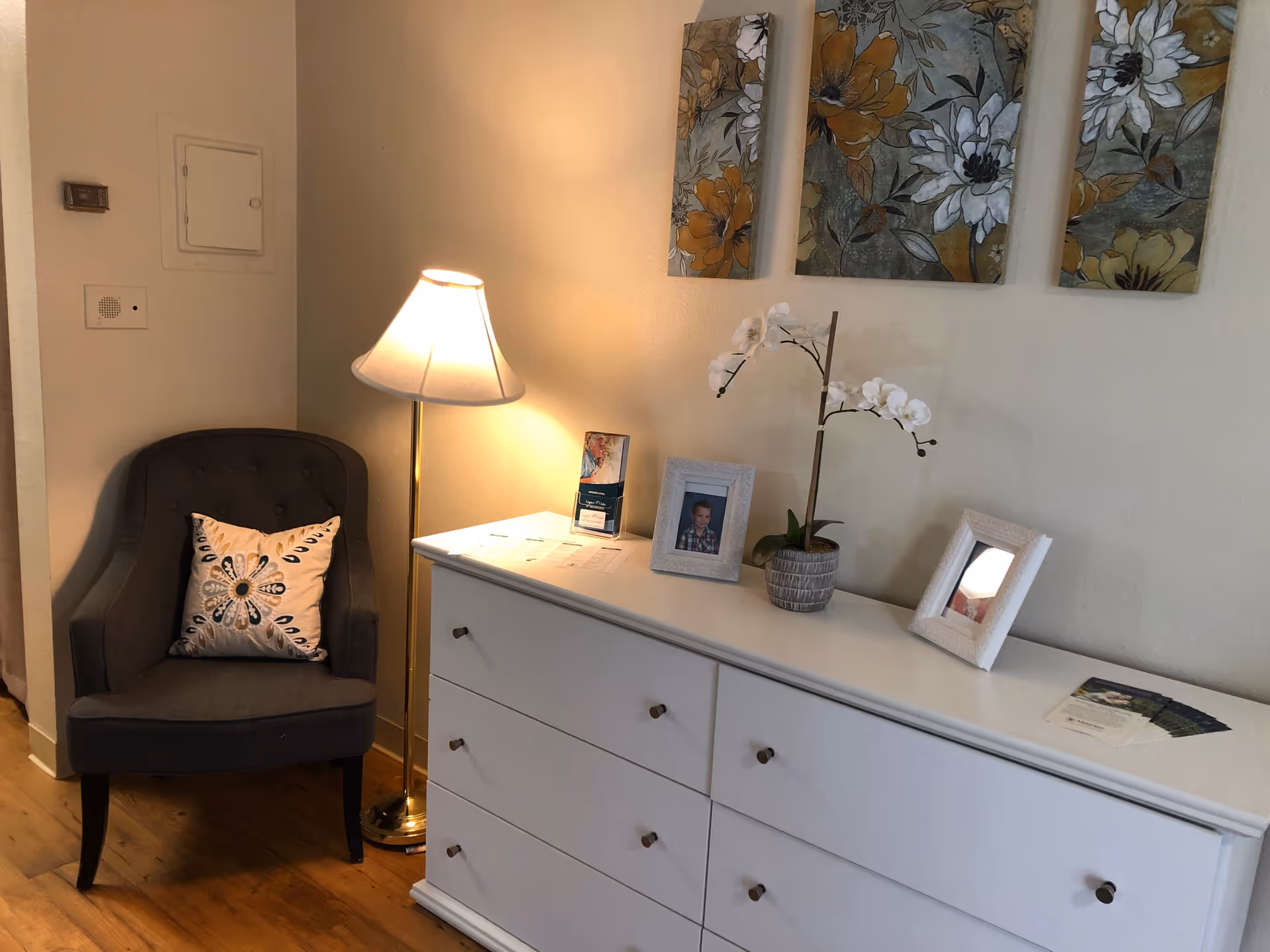 Corner of a living area with a gray armchair and floor lamp beside a white dresser topped with framed photos, a potted orchid, and floral wall art.