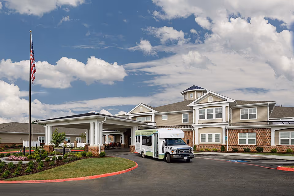 Exterior view of Bellaire at Stone Port Assisted Living & Memory Care facility showing a large building with a covered entrance, an American flag on a flagpole, landscaped greenery, and a shuttle bus parked in front under a partly cloudy sky.