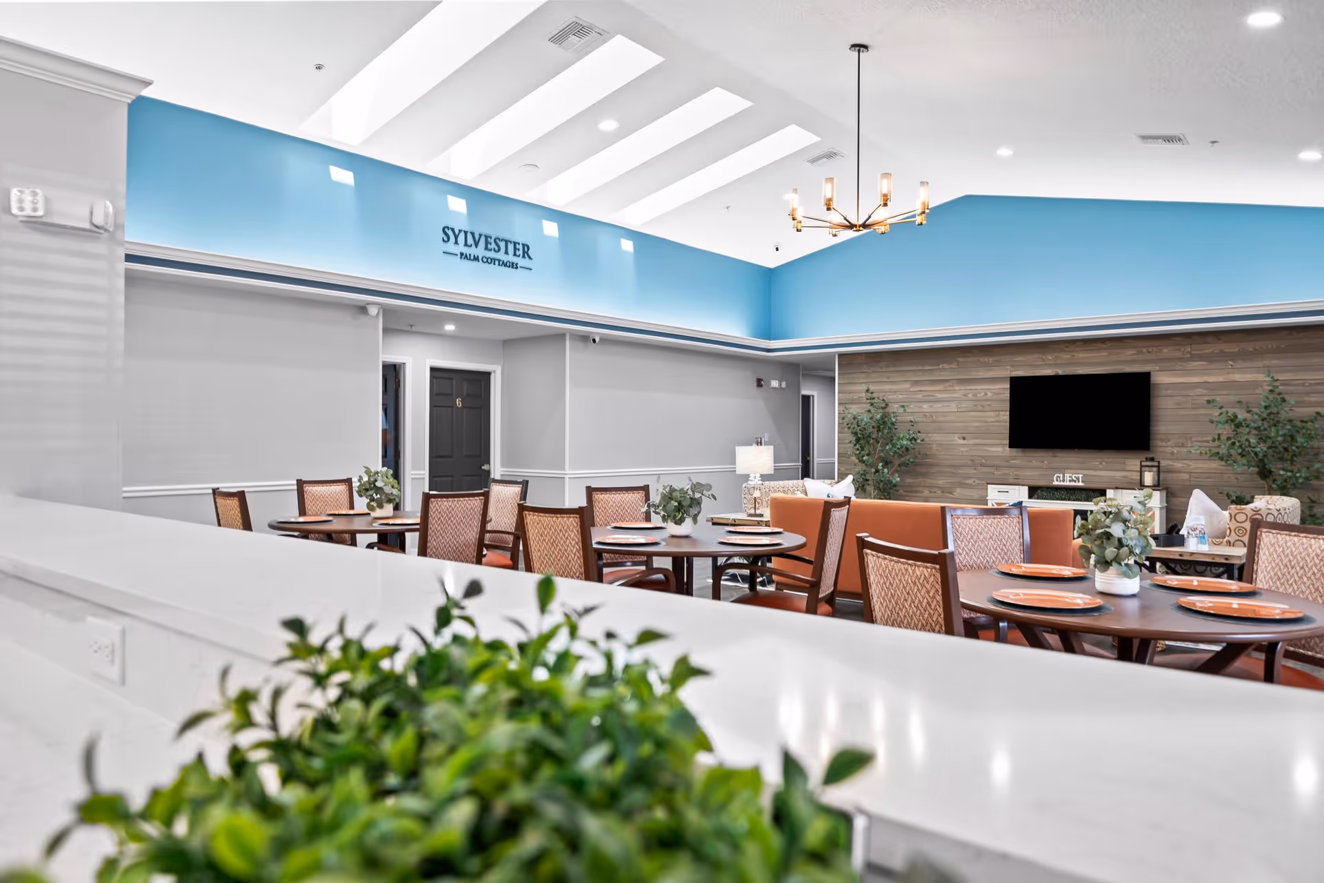 A bright and spacious common area in a senior living facility with round tables set with plates and small potted plants. The room features a high ceiling with skylights, a blue accent wall with the text 'SYLVESTER PALM COTTAGES', a flat-screen TV mounted on a wooden panel wall, and comfortable seating including chairs and sofas.