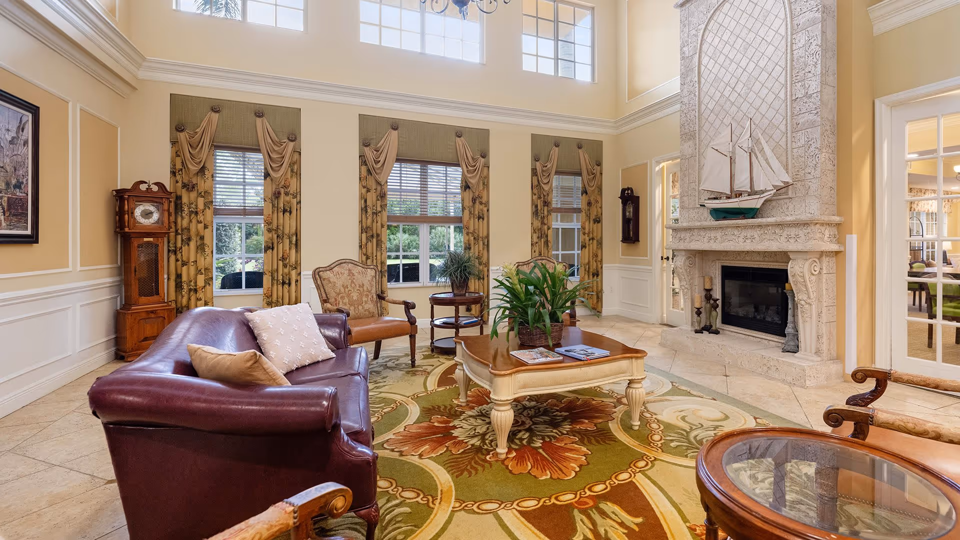 A spacious and elegant living room with high ceilings and large windows. The room features a dark leather sofa with pillows, an upholstered armchair, a wooden coffee table with plants and magazines, and a decorative fireplace with a model sailboat on the mantel. The floor is tiled and partially covered by a floral patterned area rug. There are long curtains on the windows and a grandfather clock in the corner.