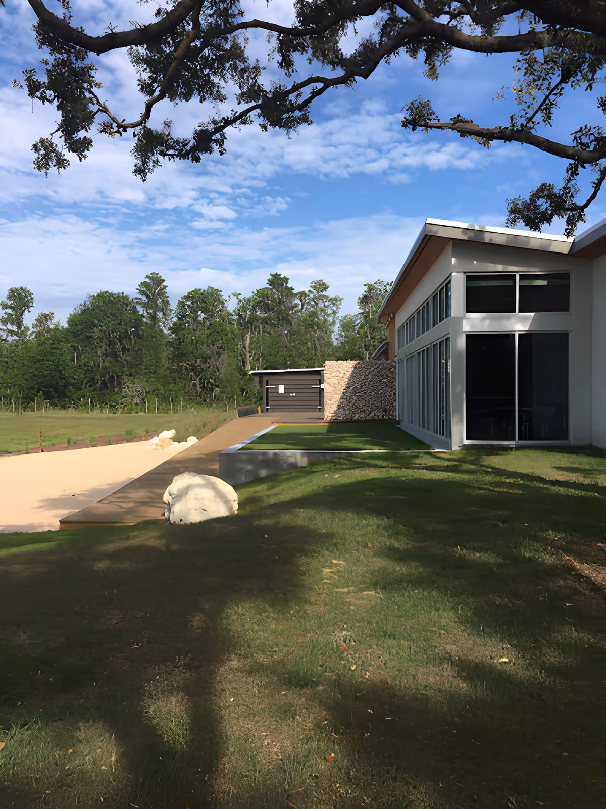 Modern single-story building with large windows beside a grassy lawn and trees under a blue sky.
