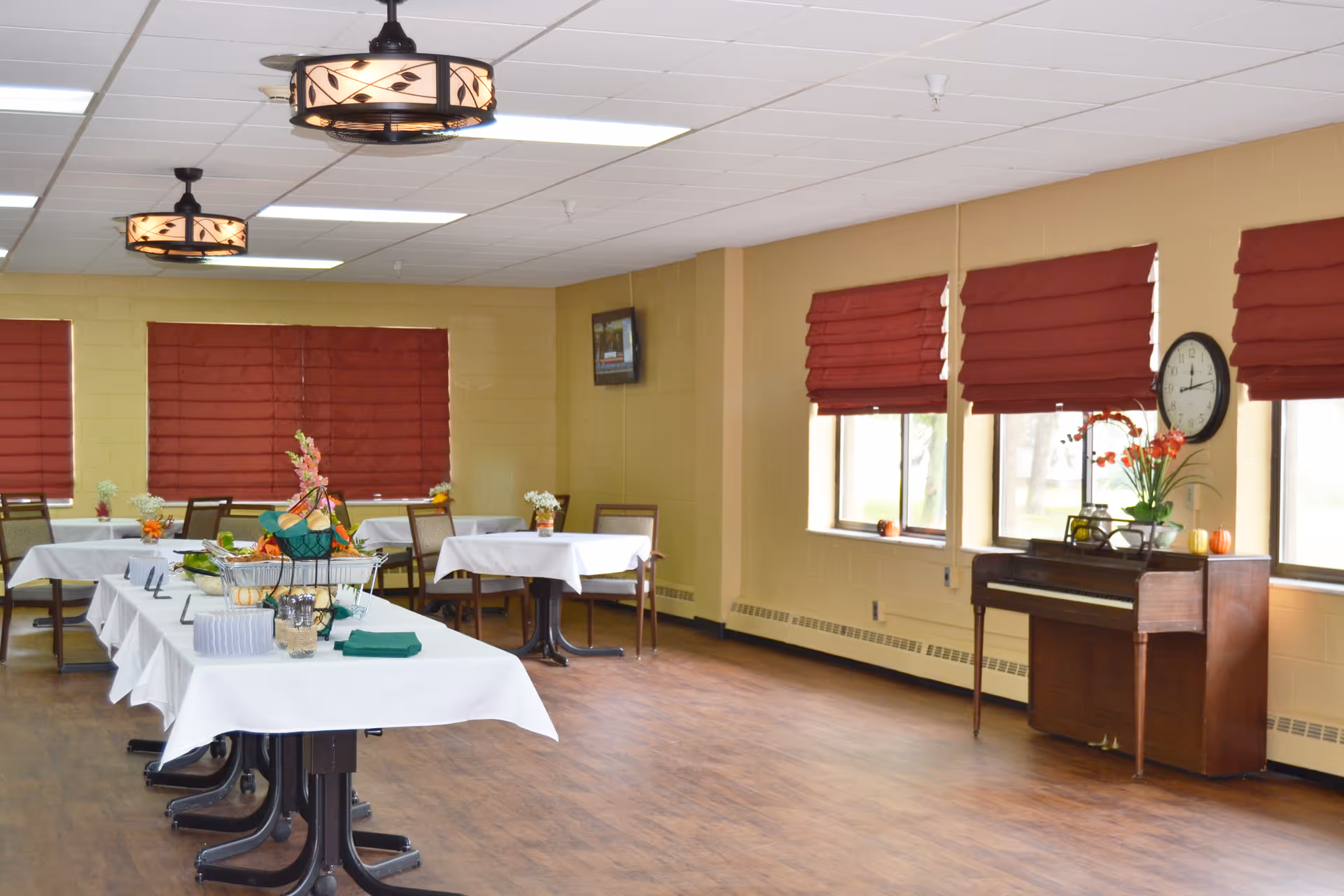 A dining room with several tables covered in white tablecloths, some decorated with small flower arrangements. The room has wooden flooring, beige walls, and windows with red blinds. A piano with a clock and a flower vase on top is positioned against one wall. Two ceiling lights with decorative shades hang from the ceiling.