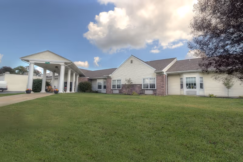Single-story retirement community building with a covered portico entrance and a large grassy front lawn under a partly cloudy sky.