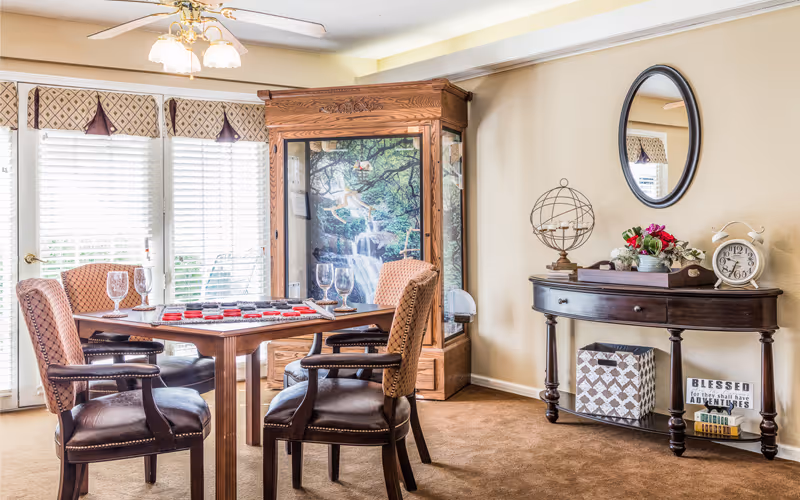 Sunlit dining/game area with a wooden table and four upholstered chairs beside a decorative console and large display cabinet.