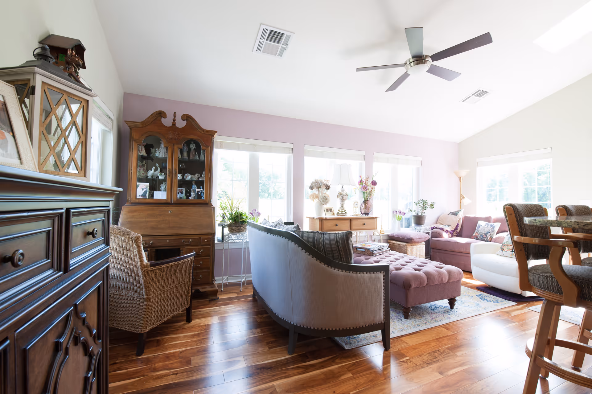 Bright and airy living room with wooden flooring, a ceiling fan, and large windows letting in natural light. The room features a mix of traditional and modern furniture including a tufted ottoman, a curved sofa, a wicker chair, a wooden cabinet with glass doors, and a dining table with chairs. Decorative items such as plants, lamps, and framed photos are placed around the room.
