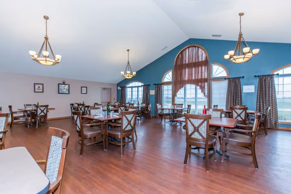 A spacious dining room with multiple wooden tables and chairs arranged neatly. The room features large arched windows with brown curtains, allowing natural light to fill the space. The walls are painted blue and beige, and there are two chandeliers hanging from the ceiling. The floor is wooden, and some tables have bottles and glasses on them.