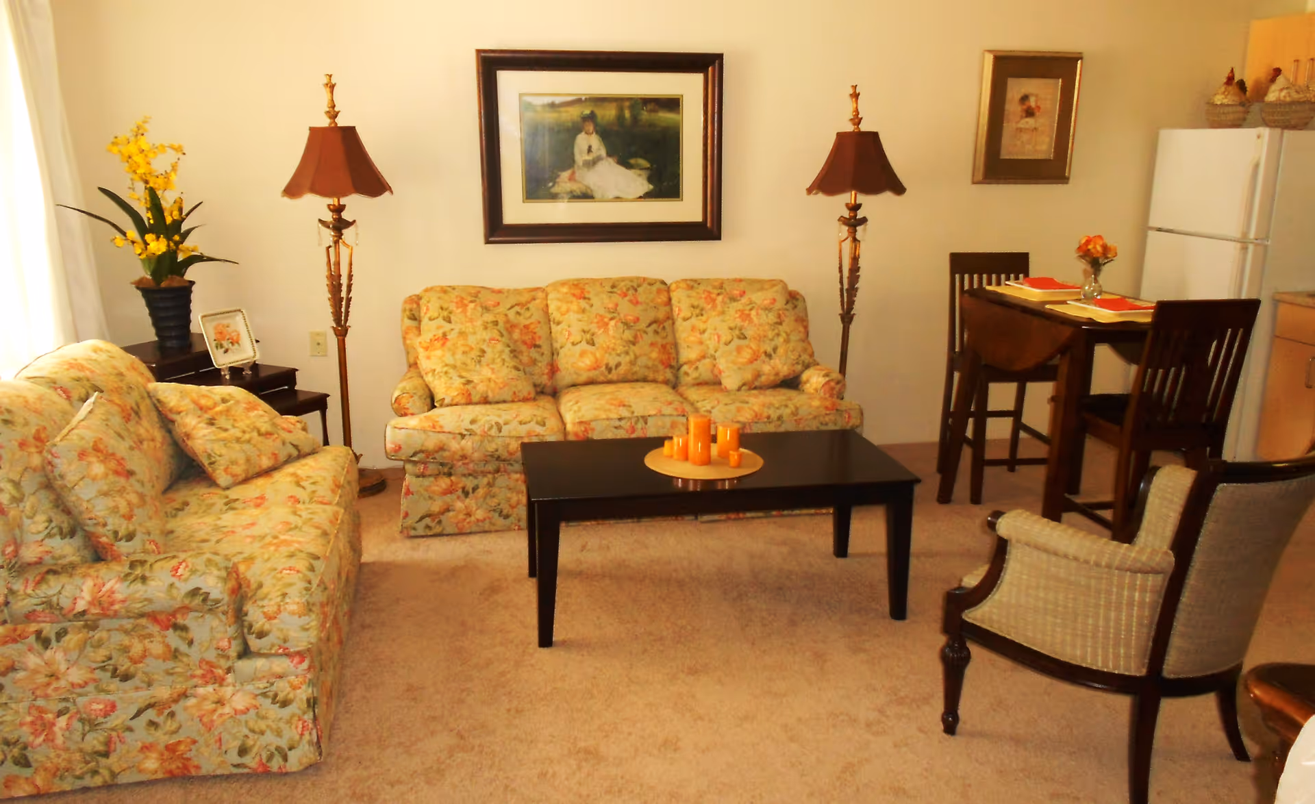 A cozy living room area with two floral patterned sofas, a dark wooden coffee table with orange candles, two tall floor lamps with brown lampshades, a small dining table with two chairs, a refrigerator, and framed artwork on the walls.