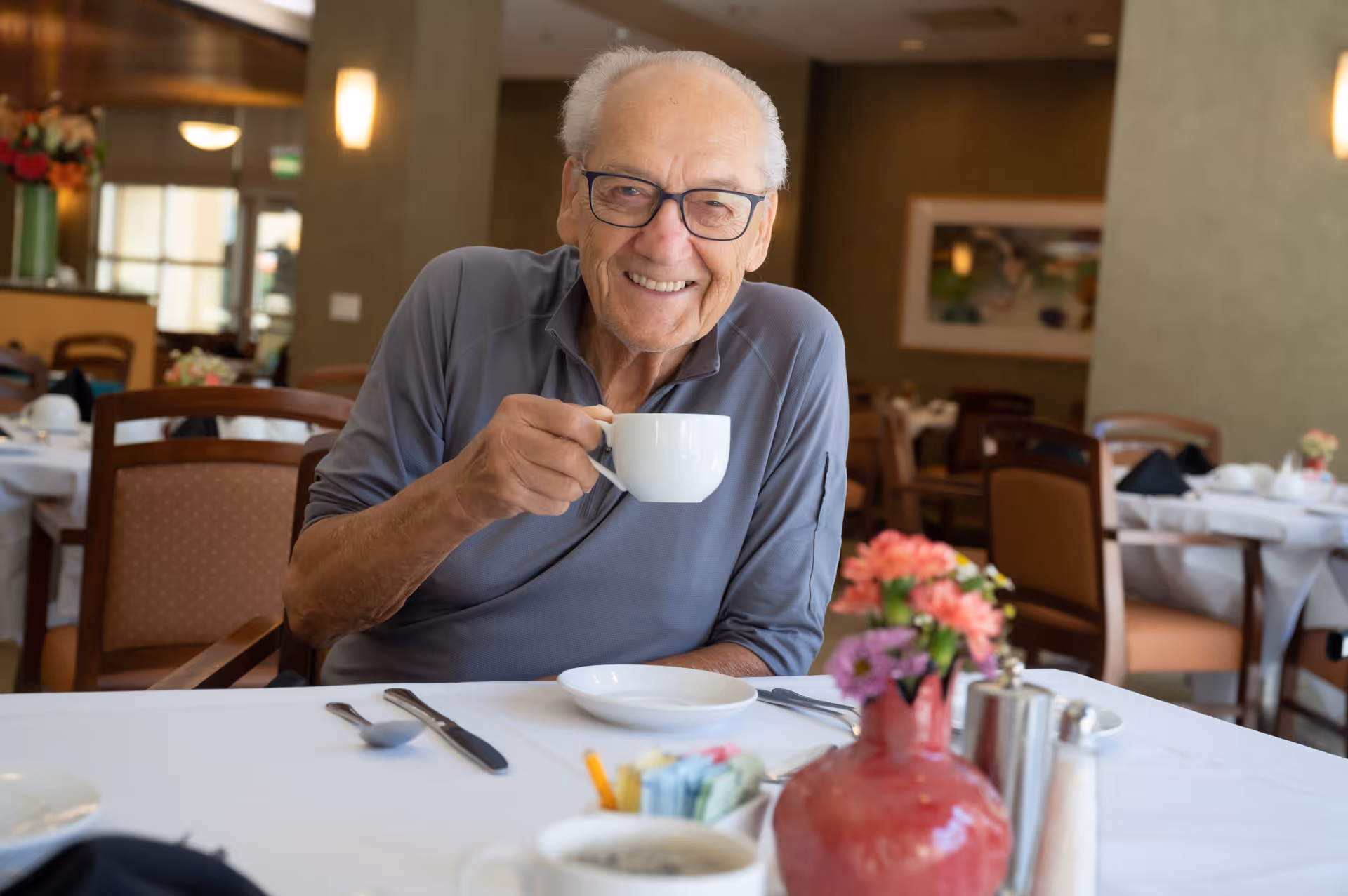 An elderly man wearing glasses and a gray shirt is sitting at a dining table in a well-lit room, smiling and holding a white cup. The table is set with utensils, a plate, a red vase with flowers, and other dining items. The background shows more tables and chairs in the dining area.