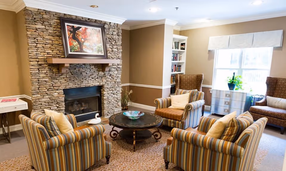 A cozy living room with a stone fireplace as the focal point. Above the fireplace is a framed painting of a tree with autumn leaves. The room has four armchairs arranged around a round coffee table with a decorative bowl. The armchairs have striped upholstery with cushions. There is a small white side table to the left and a bookshelf and window with a plant on a silver chest to the right. The walls are painted beige with white trim, and the floor is carpeted with a patterned rug.