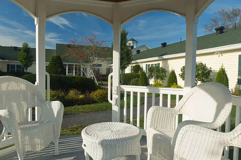White wicker chairs and a small table sit in a gazebo overlooking a landscaped courtyard and surrounding buildings.