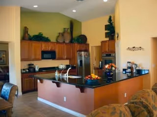 A spacious kitchen with wooden cabinets, black countertops, a stainless steel refrigerator, built-in microwave, and oven. The kitchen island has a sink and a bowl of fruit on it. Decorative vases and plants are placed on top of the cabinets. The walls are painted in warm tones of yellow and green.