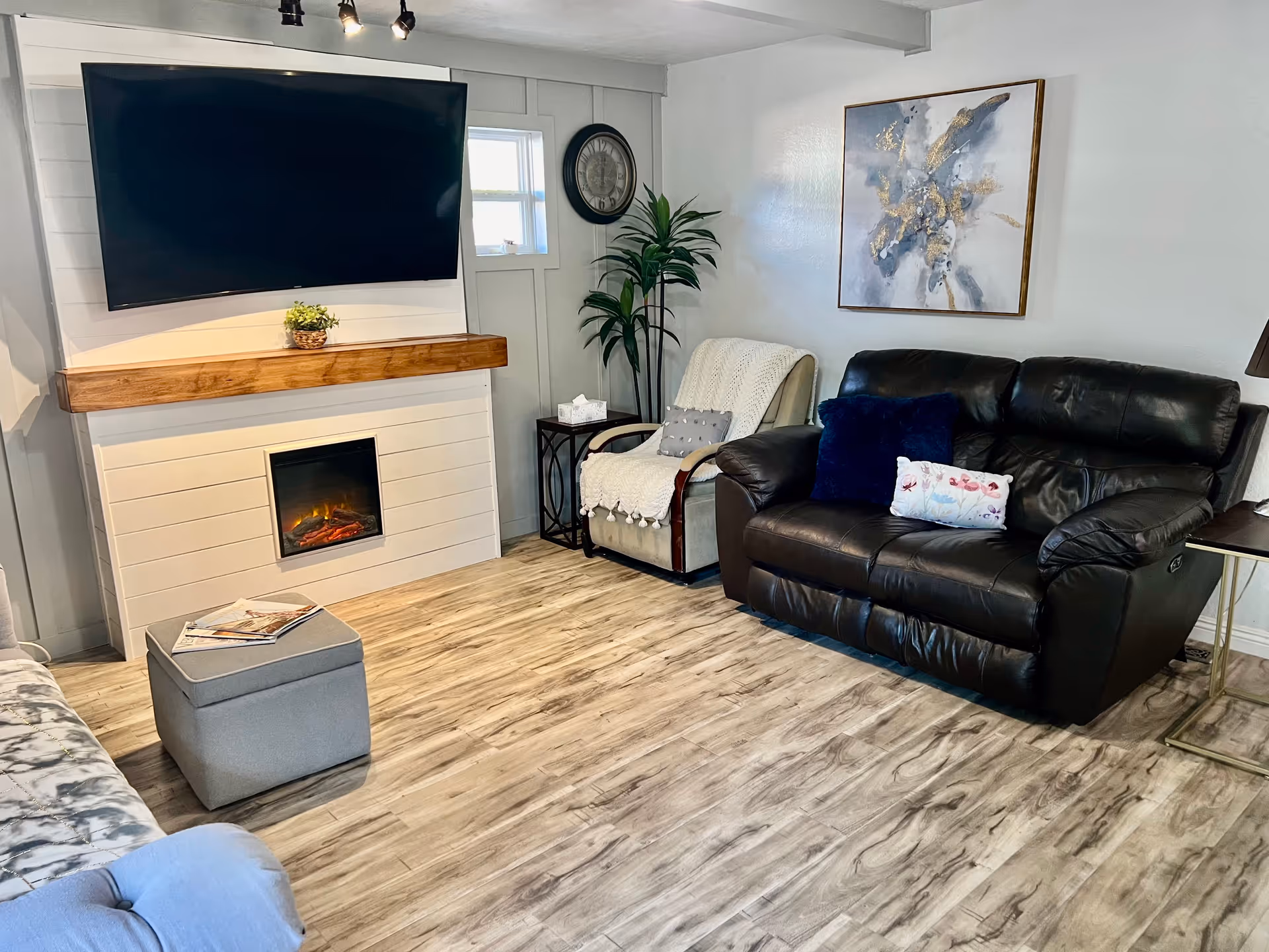 A cozy living room with a mounted flat-screen TV above a modern electric fireplace. The room features a dark leather loveseat with decorative pillows, a beige armchair with a throw blanket, a small side table with a tissue box, a potted plant, a wall clock, and a framed abstract painting. The floor is wood-patterned, and there is a gray ottoman with magazines on top.