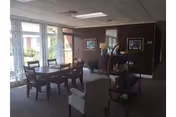 A well-lit common area in a retirement community featuring a wooden dining table with six chairs near large windows with curtains. The room has carpeted floors, framed artwork on the walls, and a small shelving unit with decorative items. There is also a comfortable armchair in the foreground.