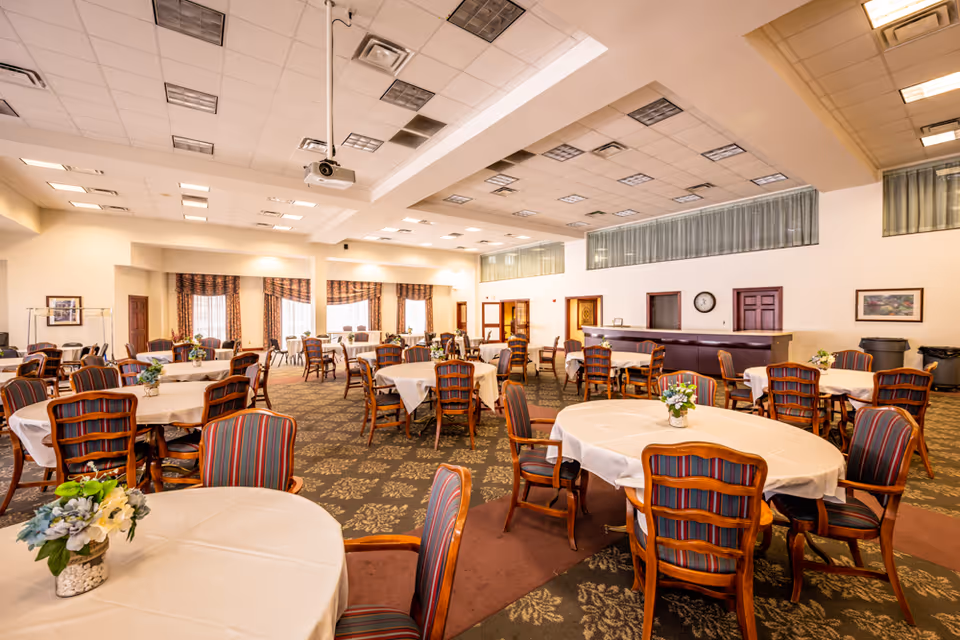 A spacious dining room with multiple round tables covered with white tablecloths and surrounded by wooden chairs with striped upholstery. Each table has a small floral centerpiece. The room has a patterned carpet, large windows with curtains, and a ceiling with numerous recessed lights and a mounted projector.